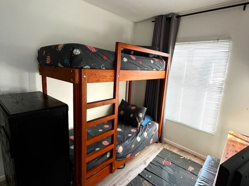A functional bedroom featuring light-toned laminate flooring, a window with venetian blinds, and a ceiling light fixture.