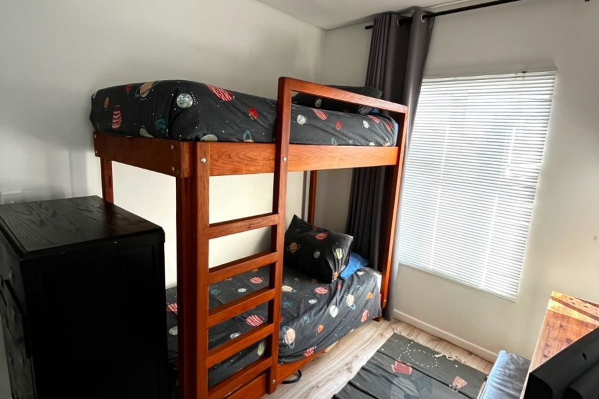 A functional bedroom featuring light-toned laminate flooring, a window with venetian blinds, and a ceiling light fixture.
