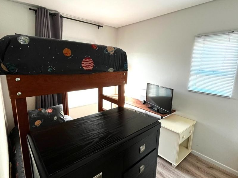 A functional bedroom featuring light-toned laminate flooring, a window with fitted venetian blinds, and a simple flush mount ceiling light fixture.
