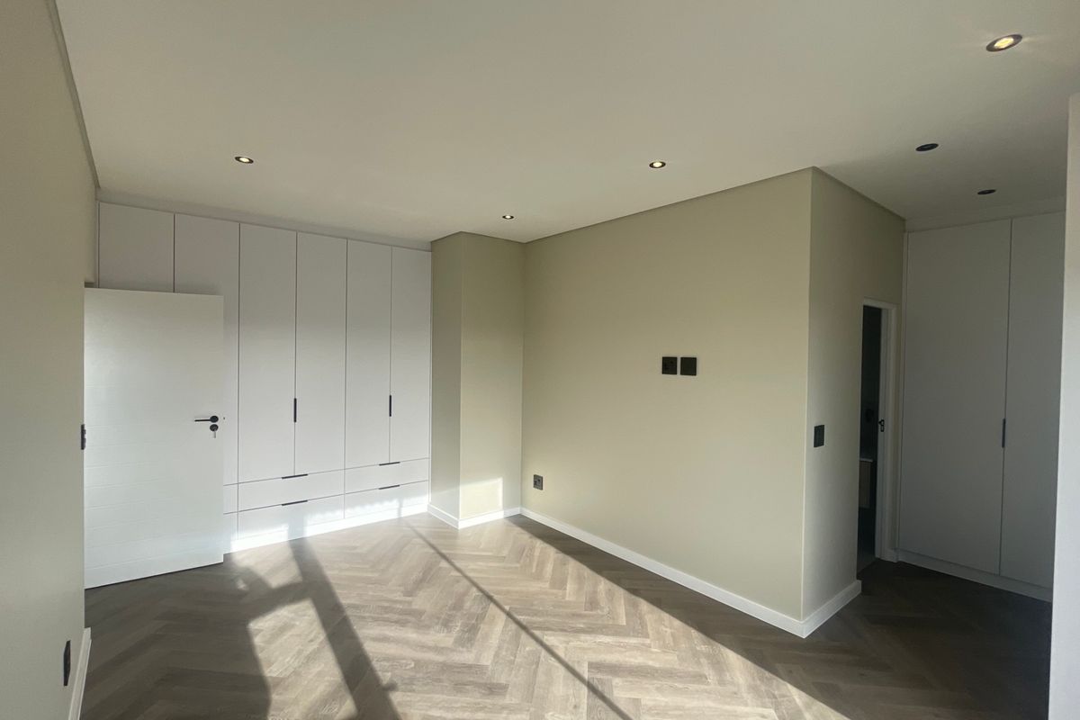 A bright, modern bedroom featuring high-quality herringbone flooring, extensive built-in cabinetry, and recessed ceiling lighting in a neutral, contemporary finish.