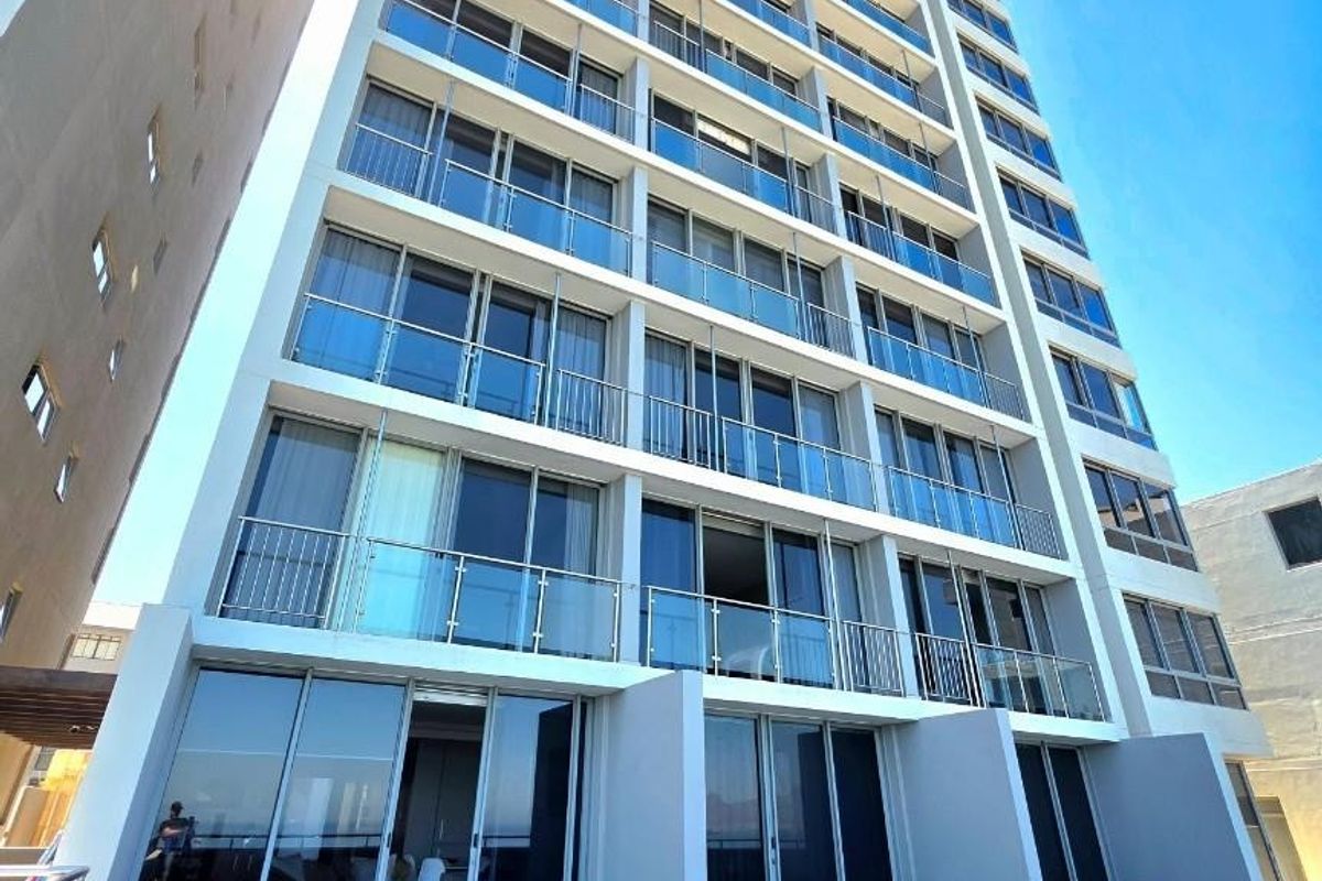 A low-angle exterior view of a modern high-rise apartment building featuring glass balconies and a paved ground-level terrace area.