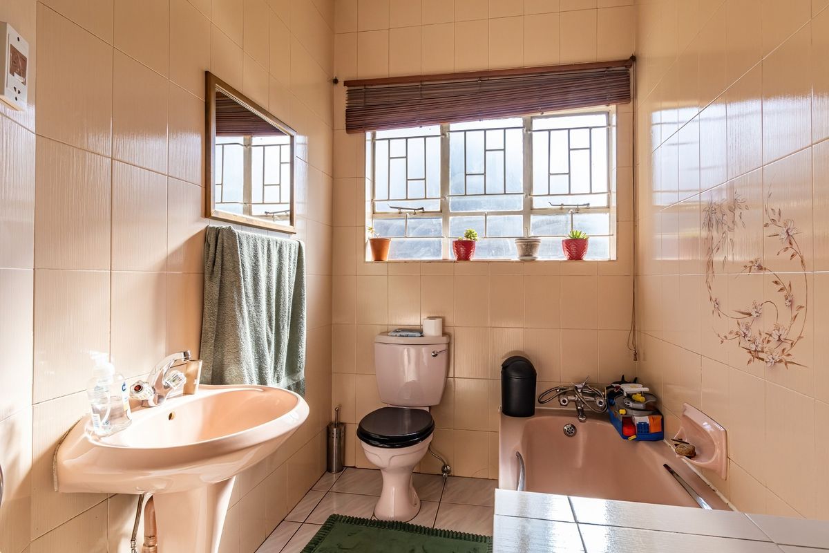 A dated bathroom featuring a peach-colored pedestal sink and bathtub, full-height wall tiling, and a window with security bars.