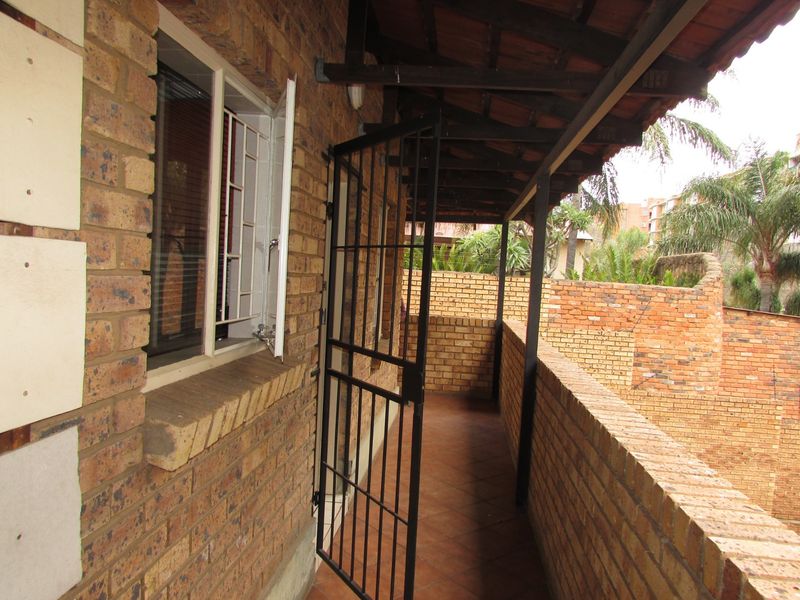 A covered brick balcony featuring a security gate, tiled flooring, and exposed timber roof structure, overlooking the surrounding complex grounds.