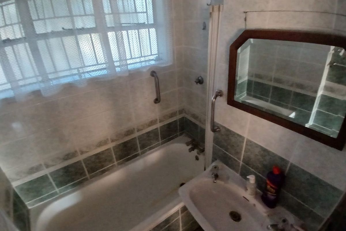 A dated bathroom featuring a built-in bathtub, wall-mounted basin, and safety grab rails, finished with older style ceramic wall tiles.