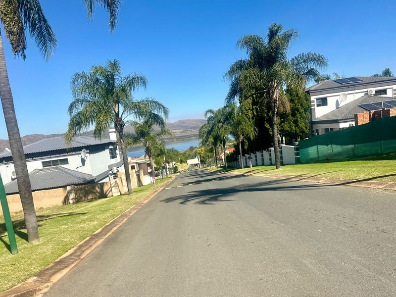 A view looking down a paved residential street in a well-maintained estate, featuring landscaped verges, palm trees, and surrounding residential properties.