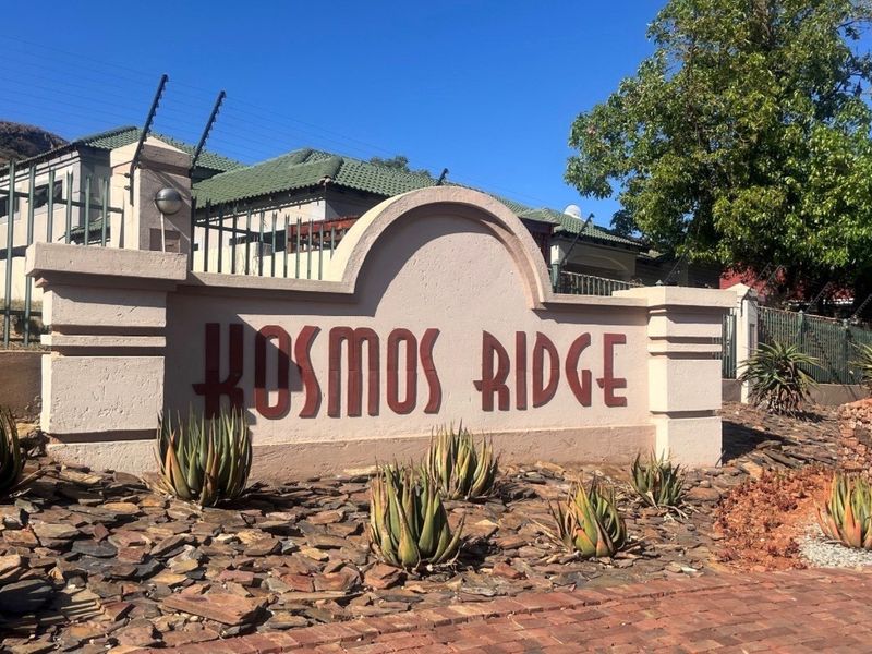 Entrance signage for the Kosmos Ridge residential estate, featuring landscaped rock garden beds and perimeter security fencing.