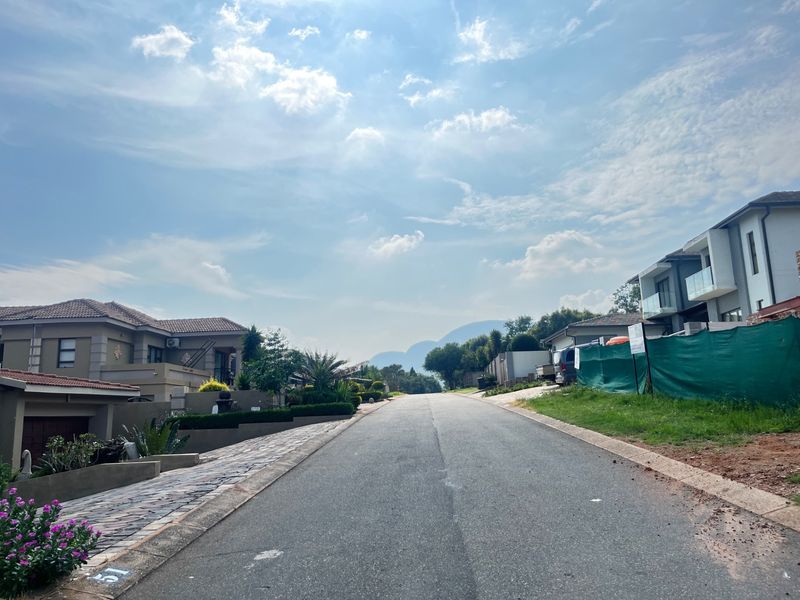 A view looking down a paved residential street in a developed neighborhood, featuring surrounding modern homes and distant mountain views.