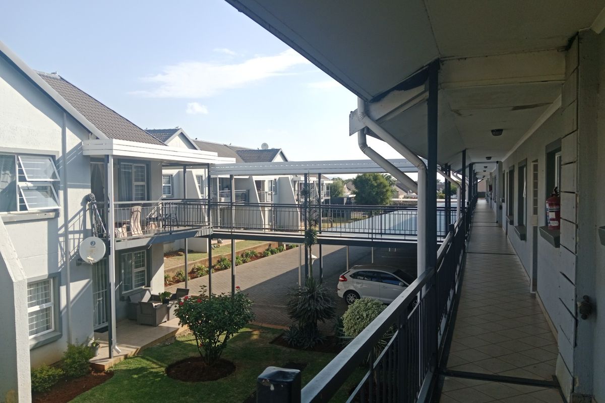 An elevated exterior walkway overlooking a landscaped courtyard and parking area within a townhouse complex, featuring tiled flooring and metal railings.
