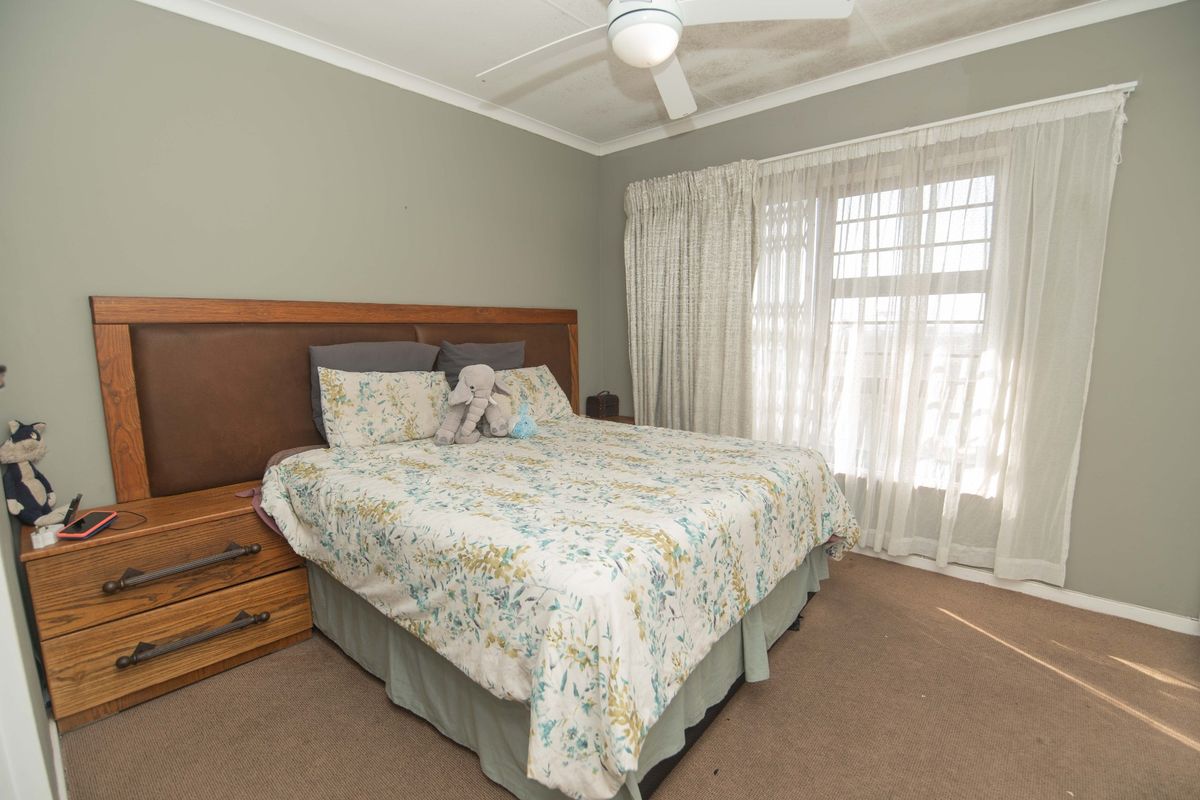 A bedroom featuring a built-in wooden headboard, ceiling fan, and large window with security gate, finished with neutral wall paint and carpet flooring.