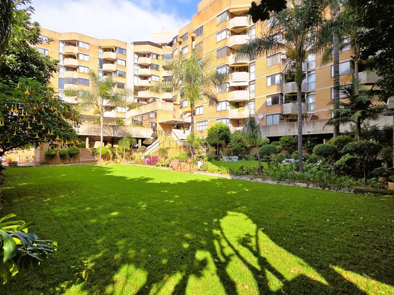A well-maintained communal garden area featuring lush green lawns and mature trees, set against the backdrop of a multi-story brick apartment building.