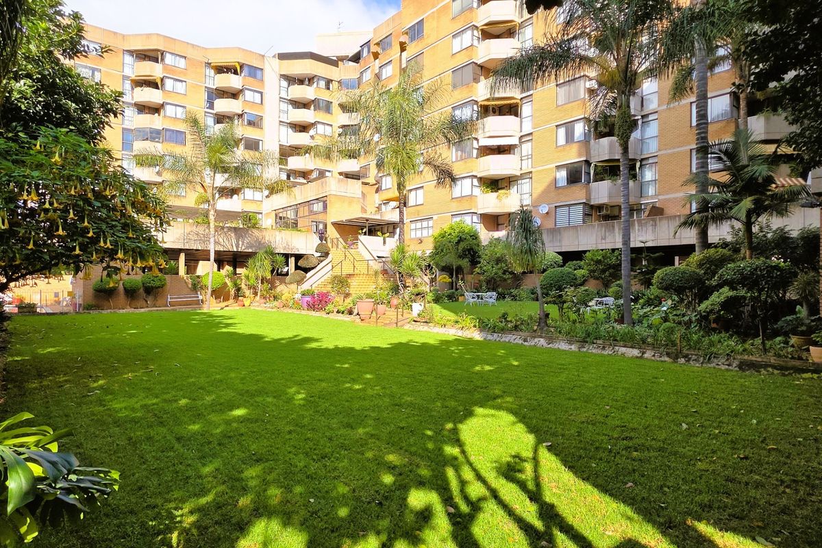 A well-maintained communal garden area featuring lush green lawns and mature trees, set against the backdrop of a multi-story brick apartment building.