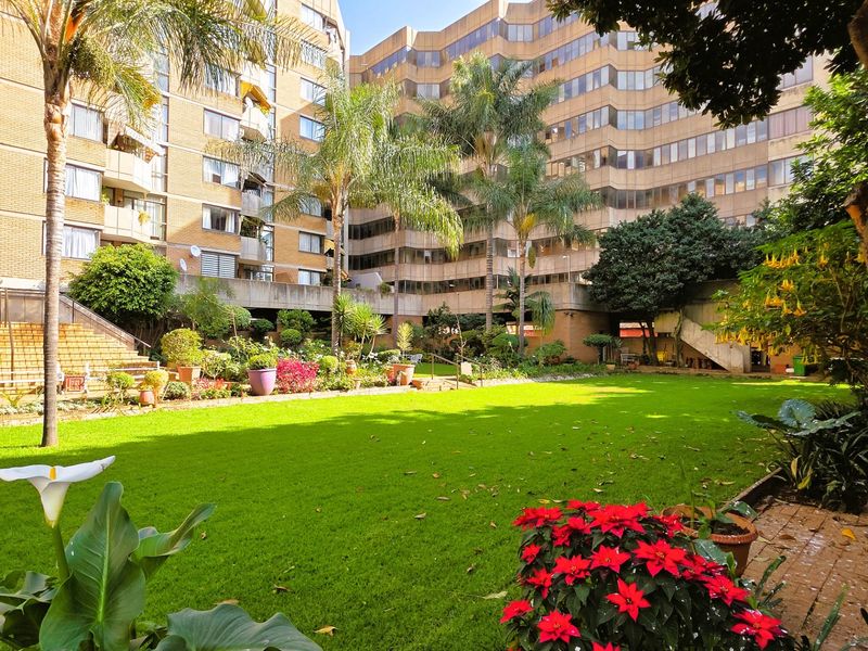 A well-maintained communal garden area featuring a lush green lawn, mature palm trees, and brick staircases set against the apartment building facade.