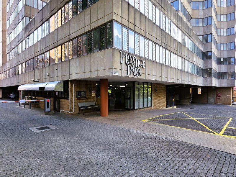 Exterior view of the Prestige Park apartment building entrance, featuring a security boom gate, paved driveway, and concrete facade.