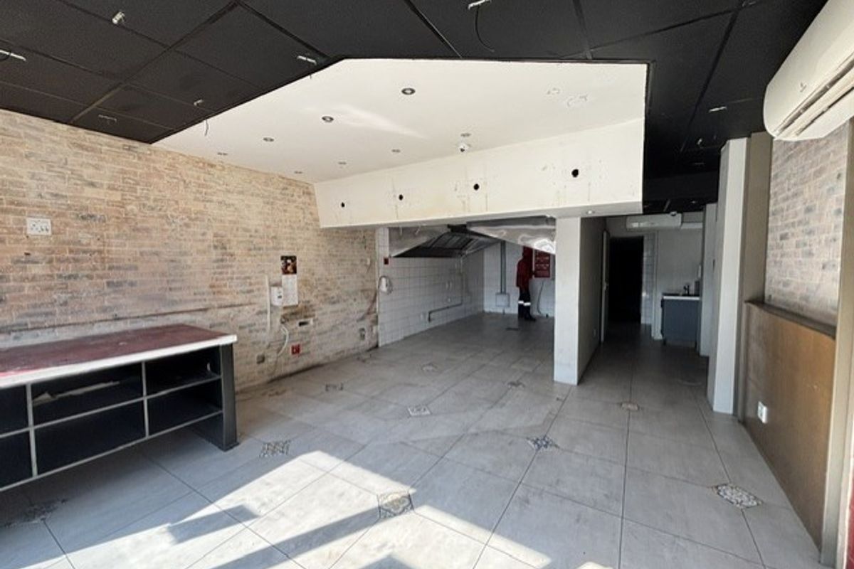 An open-plan commercial space featuring tiled flooring, exposed brick walls, a suspended ceiling, and a dedicated kitchen prep area with an extractor hood.