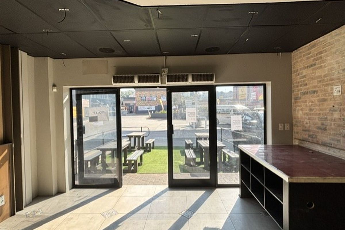 Commercial interior space featuring large glass entrance doors, tiled flooring, exposed brick wall, and overhead air conditioning unit.