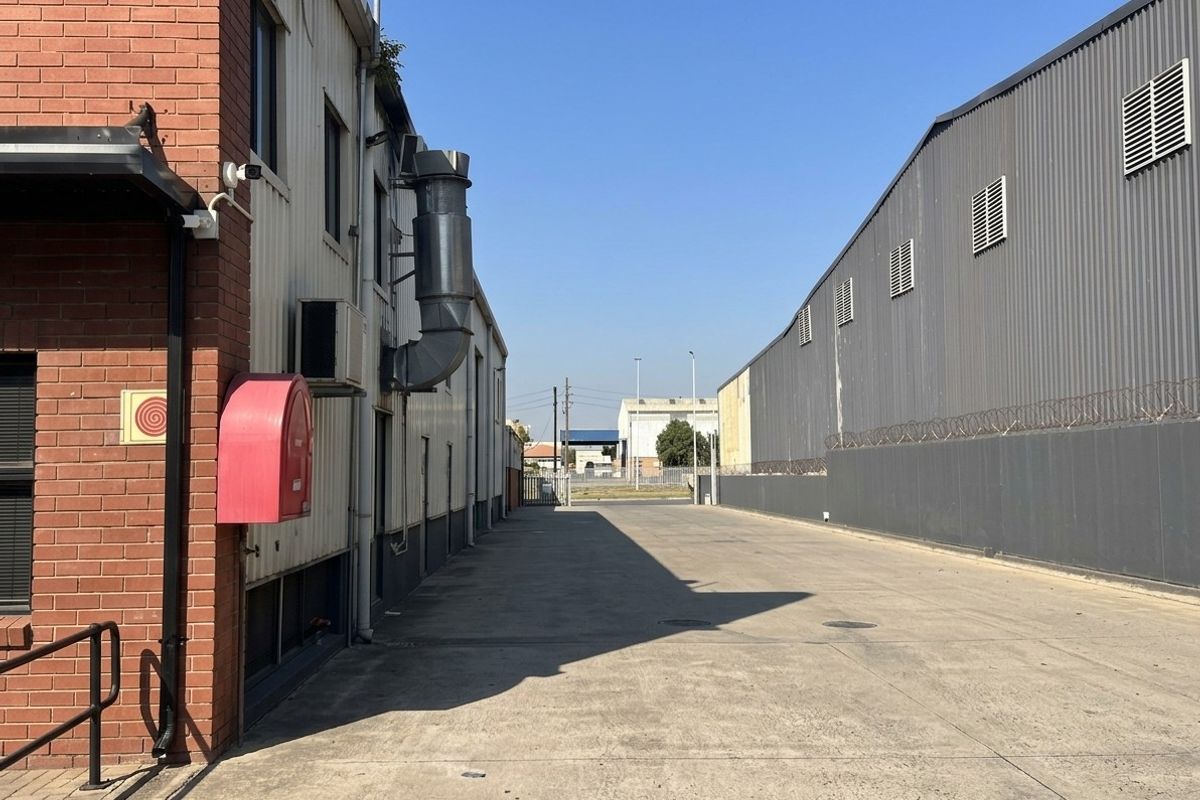 Exterior view of a commercial property showing a paved driveway between two industrial warehouse buildings with external ventilation ducting and security fencing.