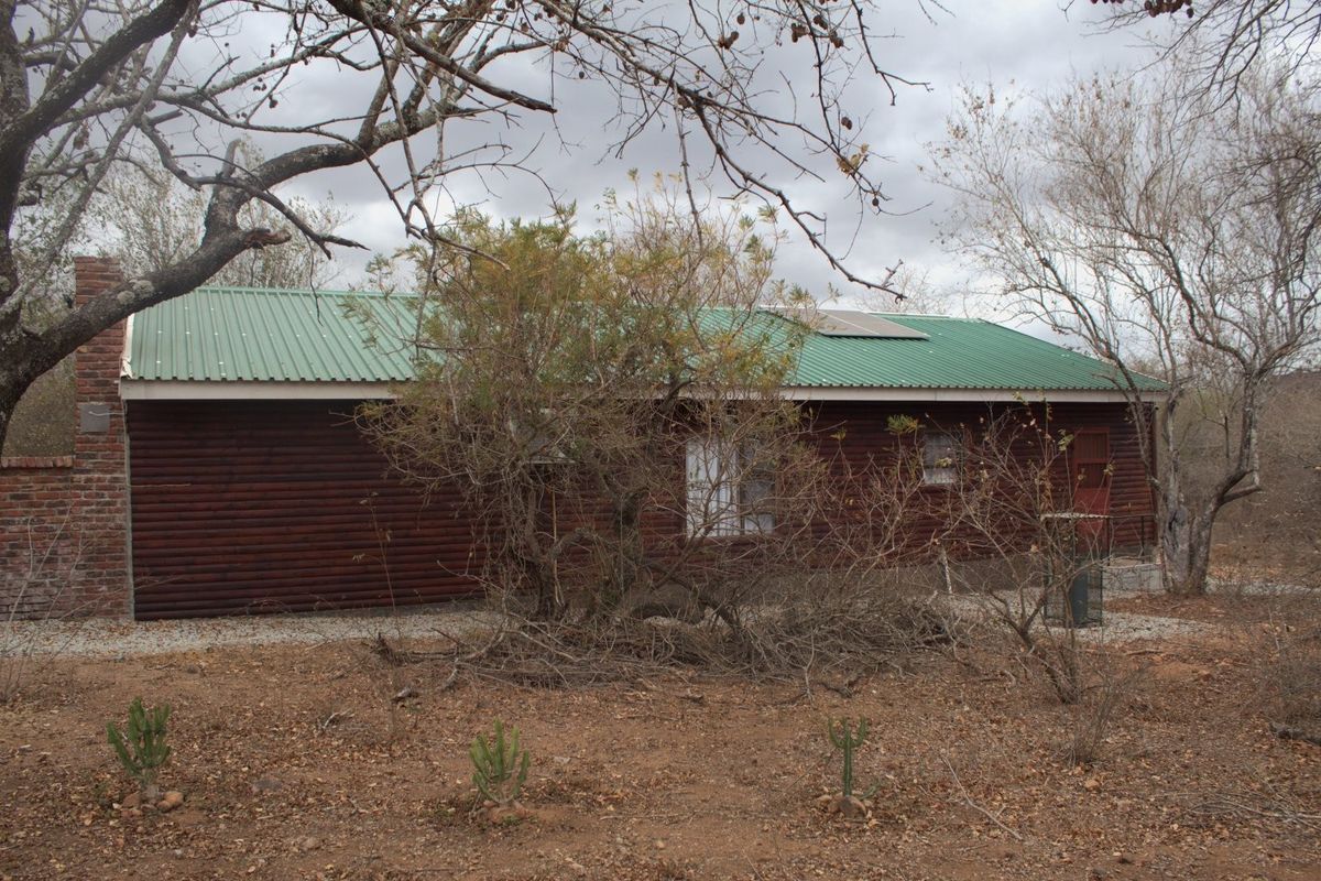 A rustic log cabin style house featuring a green corrugated metal roof, brick chimney, and solar panels, set in a natural bushveld environment.