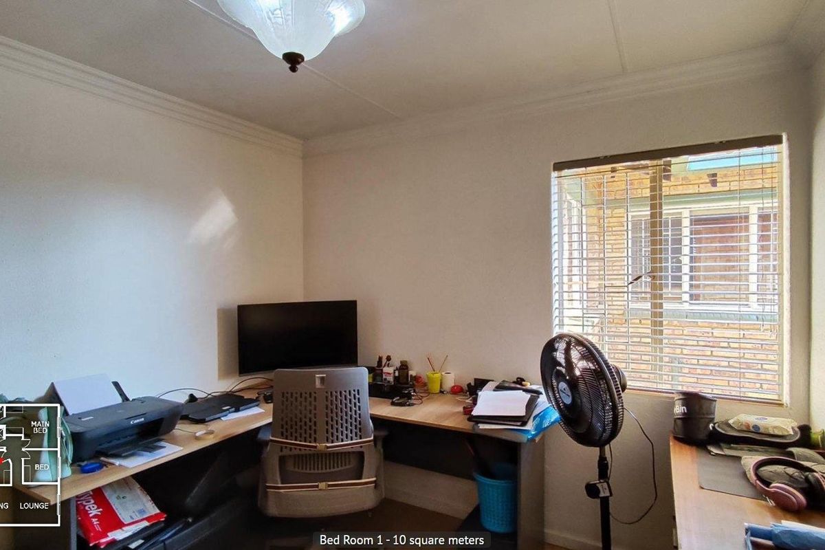 A functional bedroom currently utilized as a home office, featuring a window with fitted blinds and standard ceiling lighting.