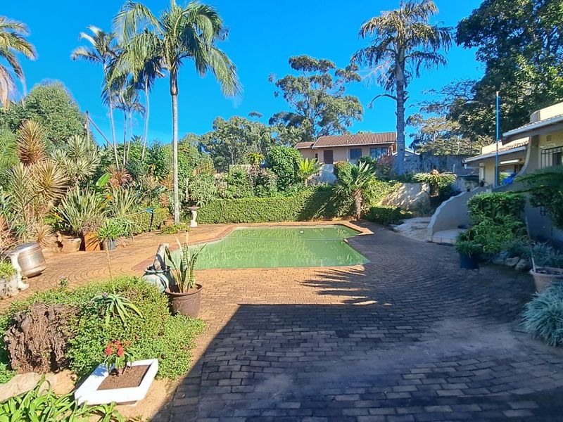 A spacious outdoor pool area featuring a swimming pool, extensive brick paving, and a lush, established garden with mature trees and shrubs.