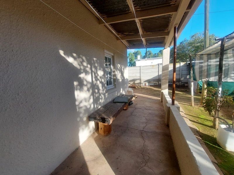 A covered outdoor patio area featuring a concrete floor, structural support pillars, and a corrugated roof, overlooking a rear garden space.