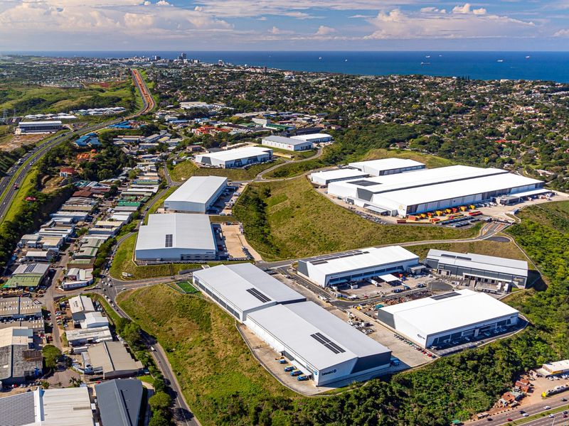 Aerial view of a large industrial park featuring multiple warehouse facilities, loading bays, and access roads, situated near a coastal urban area.