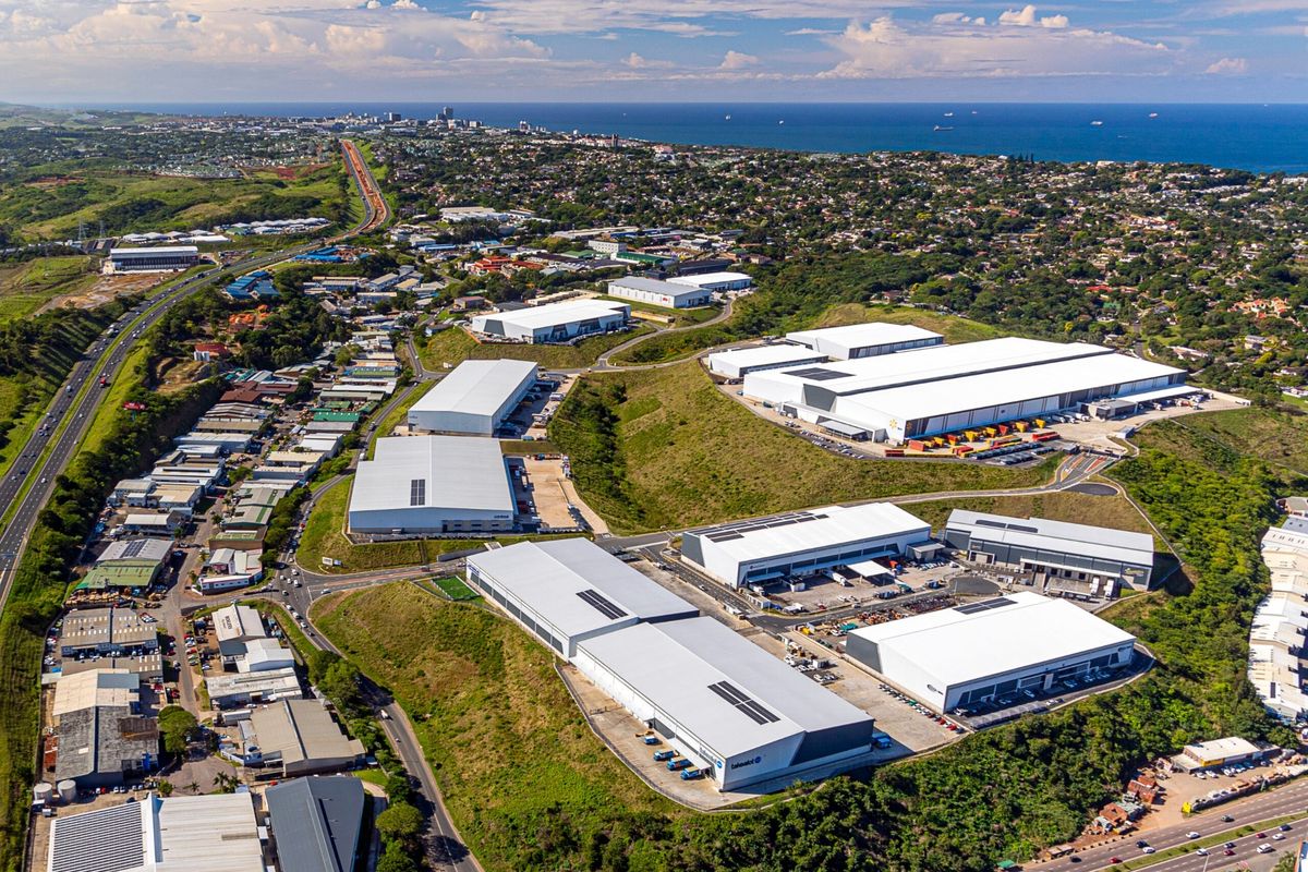 Aerial view of a large industrial park featuring multiple warehouse facilities, loading bays, and access roads, situated near a coastal urban area.
