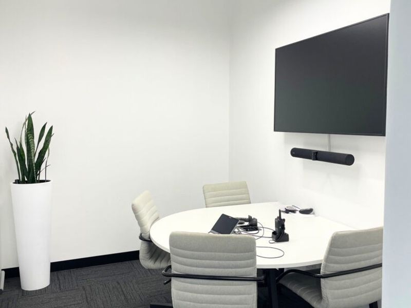 A modern, well-lit office meeting room featuring a contemporary triangular pendant light, wall-mounted display screen, and professional carpet flooring.