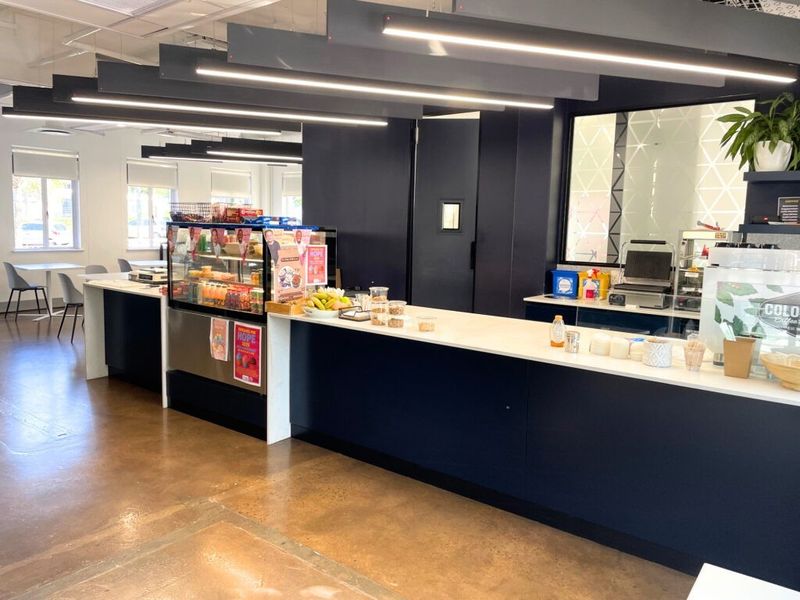 Modern office kitchenette and coffee service area featuring sleek dark cabinetry, stone countertops, and contemporary linear pendant lighting.