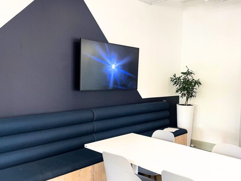 Modern office breakout area featuring built-in bench seating, a wall-mounted display screen, and an exposed industrial-style ceiling grid.