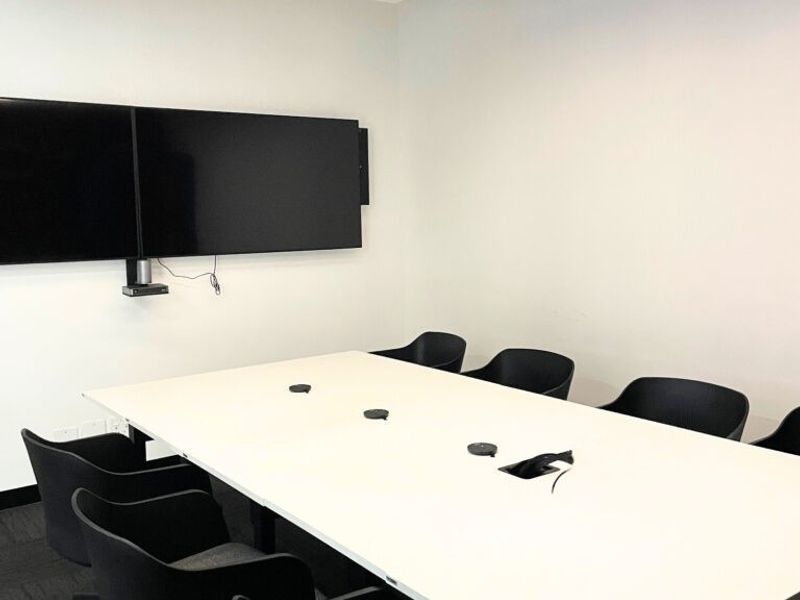 A modern, professional office meeting room featuring sleek black linear pendant lighting, dark carpeting, and a wall-mounted dual-screen display system.