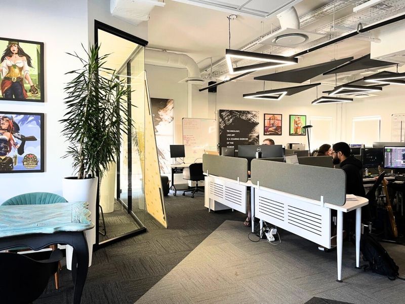 Modern open-plan office space featuring exposed industrial ceiling, contemporary geometric lighting fixtures, glass-partitioned meeting room, and carpeted flooring.