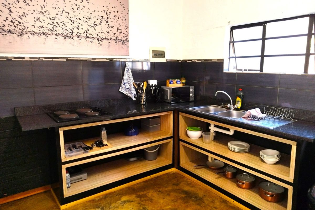 A functional kitchen area featuring dark granite countertops, open wooden shelving, a built-in electric hob, and a double stainless steel sink.
