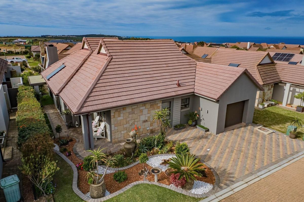 Aerial view of a well-maintained residential property featuring a tiled roof, stone cladding, paved driveway, single garage, and landscaped garden with ocean views.