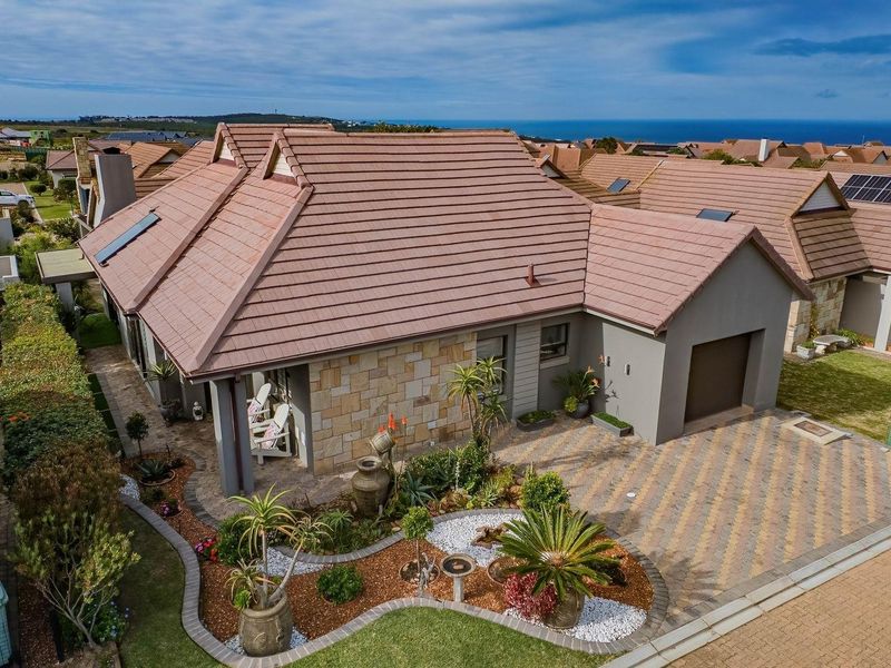 Aerial view of a well-maintained residential property featuring a tiled roof, stone cladding, paved driveway, single garage, and landscaped garden with ocean views.