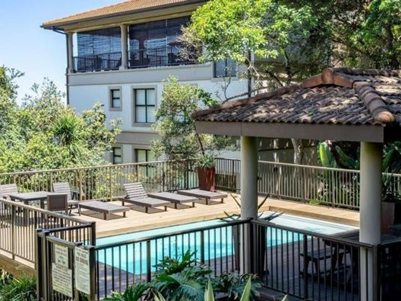 A well-maintained communal swimming pool area featuring timber decking, a covered gazebo, and secure metal balustrading, set within a landscaped apartment complex.
