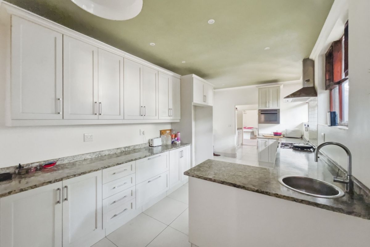 A functional kitchen featuring white cabinetry, granite countertops, a stainless steel extractor fan, and tiled flooring with a connected scullery area.