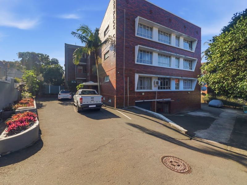 Front exterior view of a multi-story brick apartment building featuring a paved driveway, basement parking access, and established landscaping.