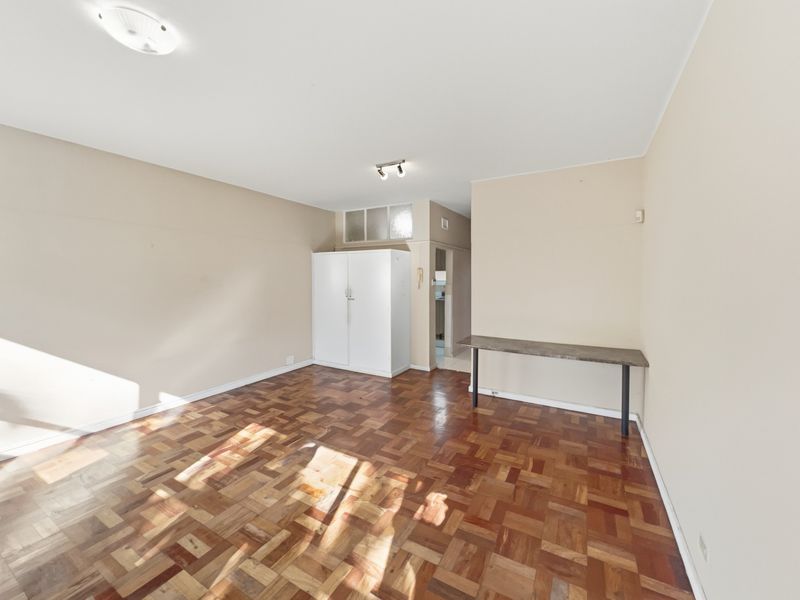 A bright, empty living room featuring classic parquet flooring, a built-in storage cupboard, and a fixed wall-mounted desk area.