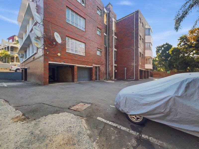 Exterior view of a multi-story brick apartment building featuring ground-level garage access and an asphalt parking area.