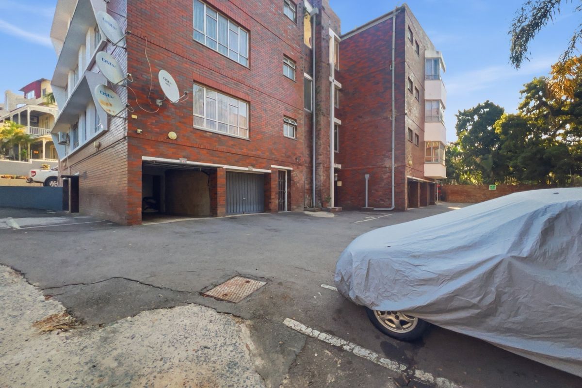 Exterior view of a multi-story brick apartment building featuring ground-level garage access and an asphalt parking area.