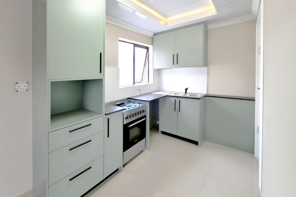A modern, recently renovated kitchen featuring light green cabinetry, a gas stove with oven, double sink, and clean tiled flooring with recessed ceiling lighting.