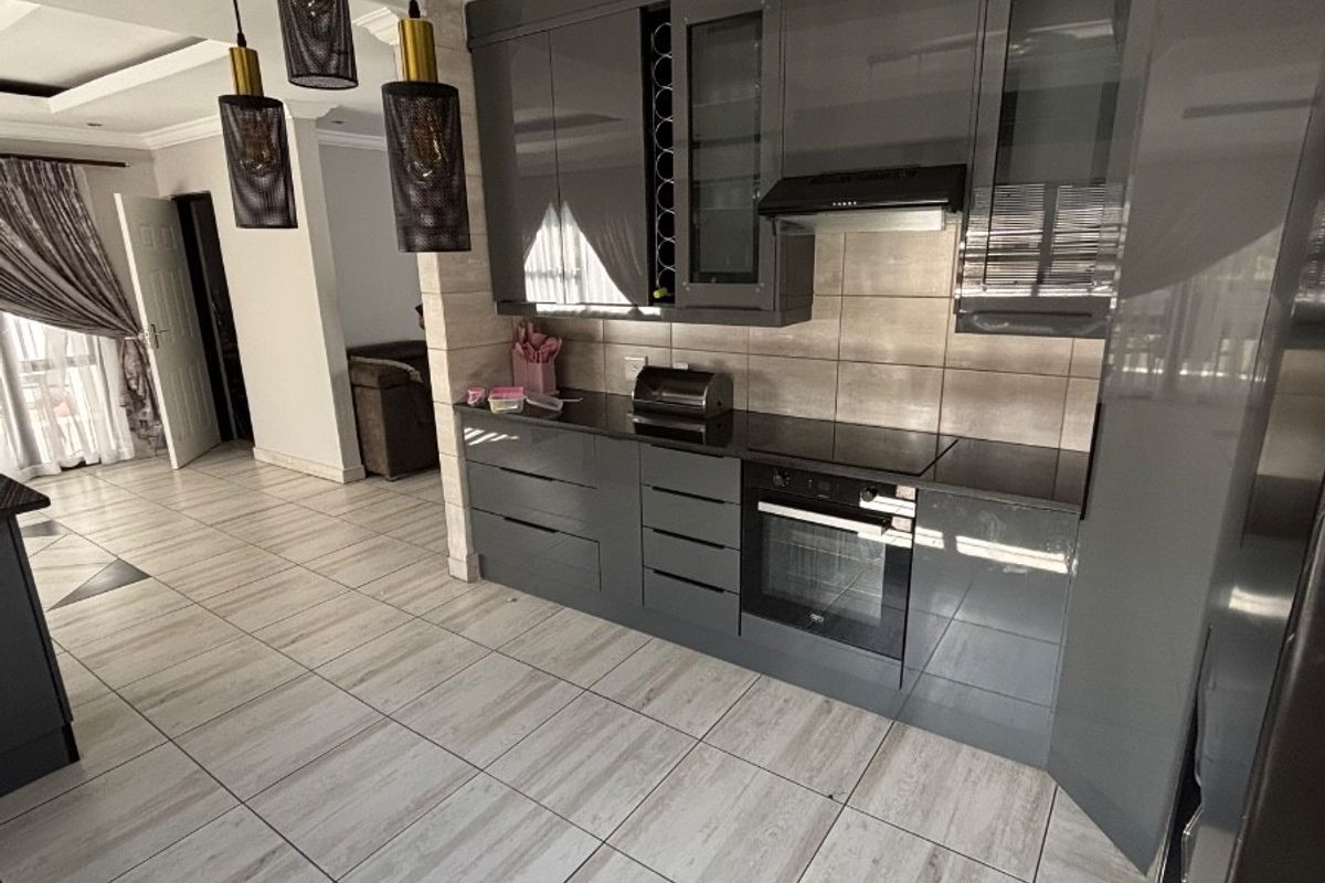 Modern kitchen featuring high-gloss grey cabinetry, integrated oven, hob, extractor fan, and tiled flooring, seamlessly flowing into the adjacent living area.