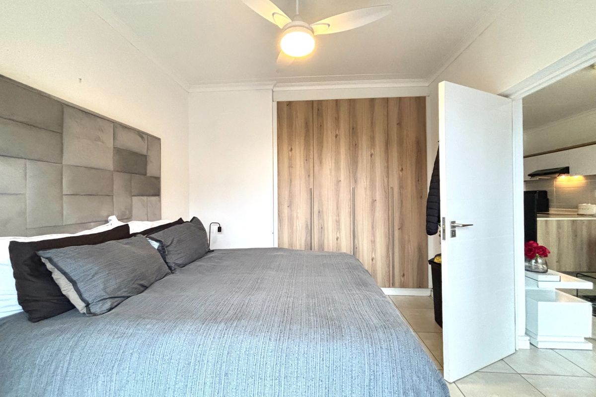A well-maintained bedroom featuring built-in wooden cabinetry, a ceiling fan, and tiled flooring, with an adjacent view into the kitchen area.