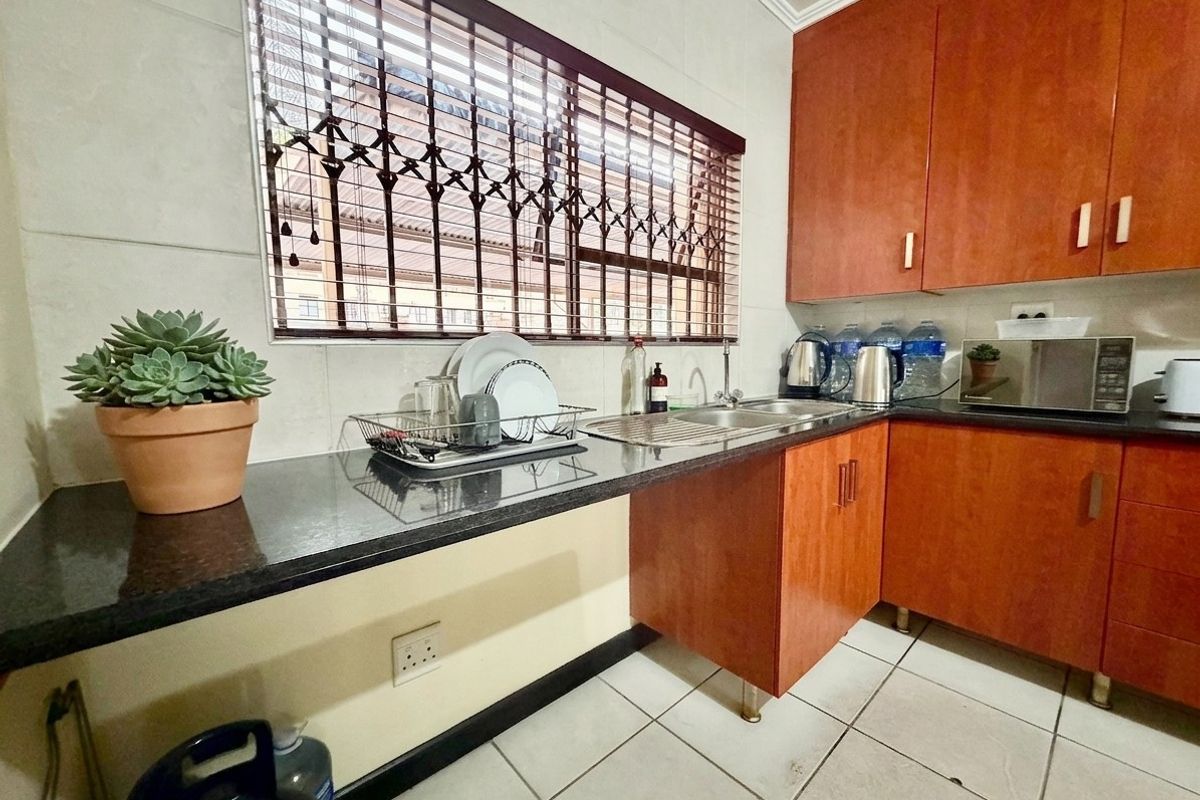 A functional kitchen featuring wooden cabinetry, granite countertops, a stainless steel sink, and tiled flooring, with security bars installed on the window.