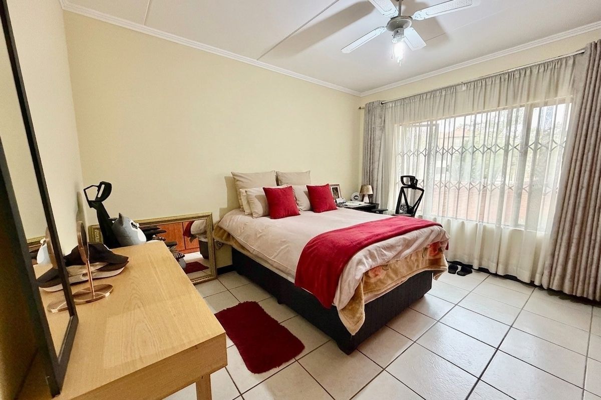 A functional bedroom featuring tiled flooring, a ceiling fan, and a large window fitted with a security gate and curtains.