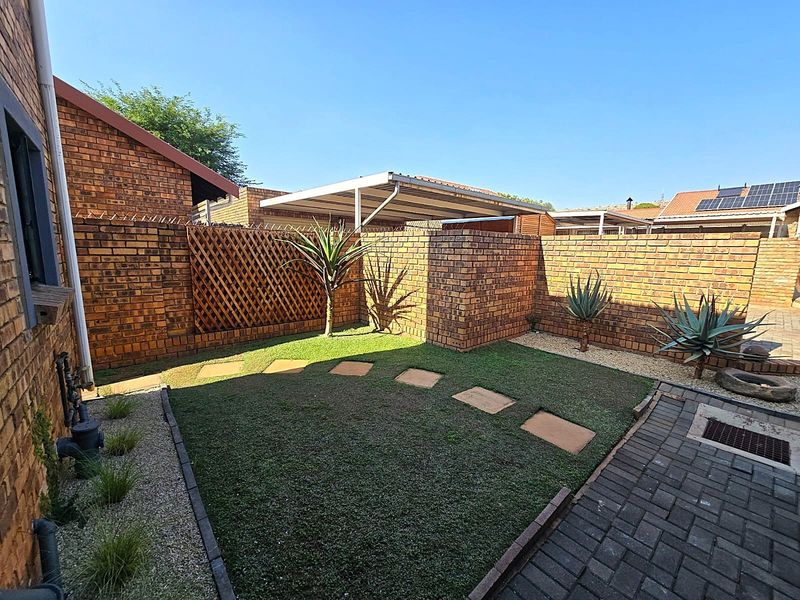 A well-maintained rear garden featuring a brick boundary wall, paved walkway, artificial lawn, and a covered carport structure.