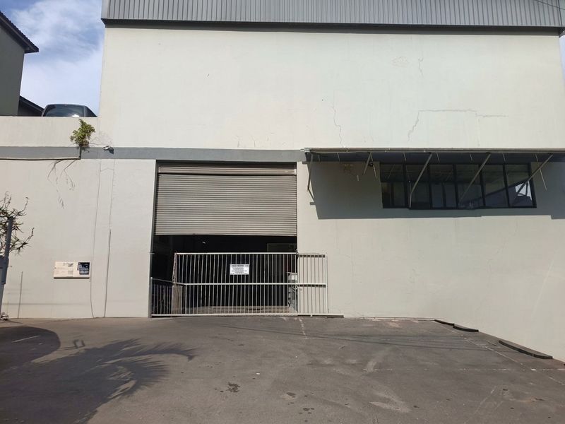 Industrial property exterior featuring a roller shutter door, security gate, and asphalt driveway, showing signs of wear and maintenance requirements.