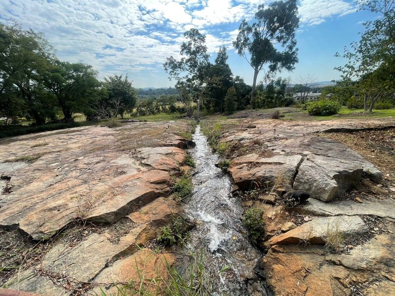 A scenic view of a natural rocky landscape featuring a flowing stream, surrounded by mature trees and open greenery.