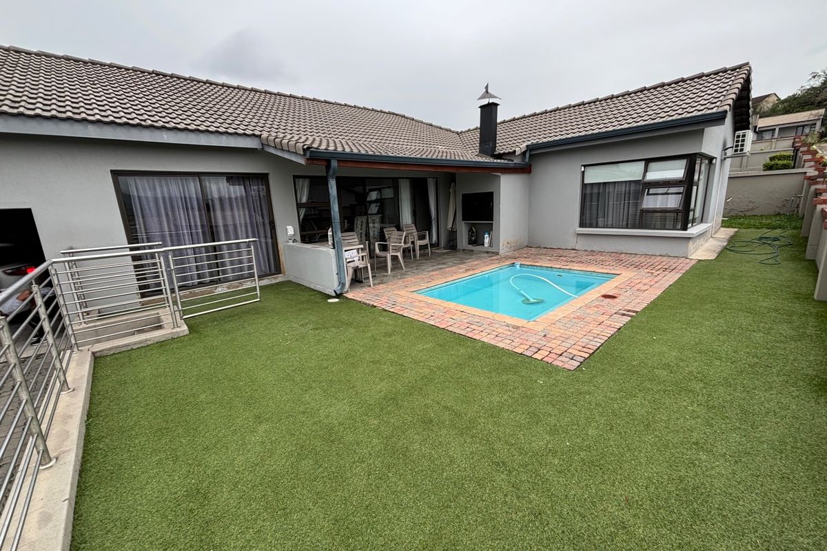Rear exterior view featuring a small swimming pool with brick surround, artificial lawn, and a covered patio area with an outdoor fireplace.