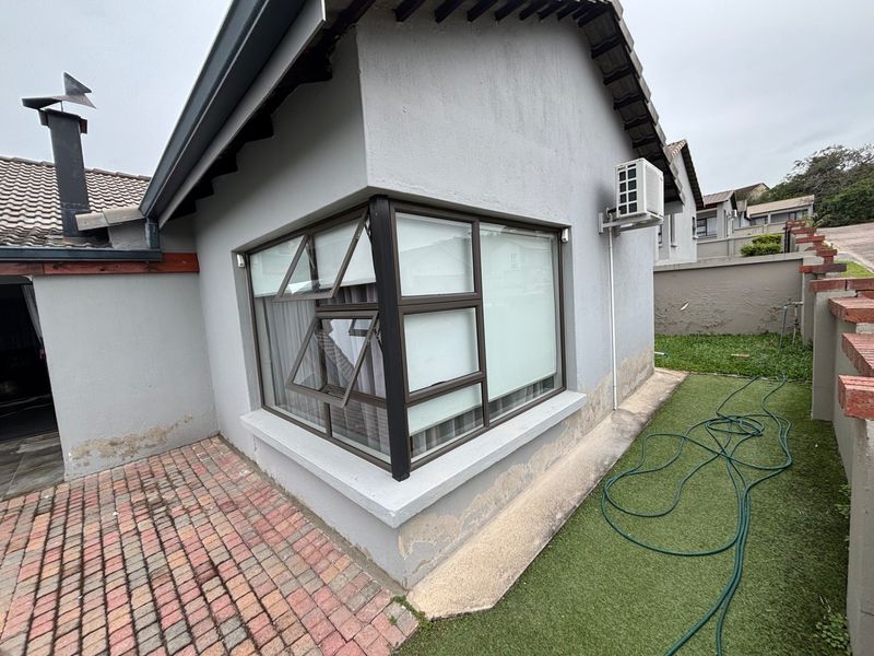 Rear exterior view of the property featuring a corner window, wall-mounted air conditioning unit, brick paved patio area, and artificial lawn.